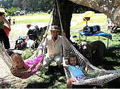 Two young girls relax in rope hammocks beneath a large shade tree. Their father, a man in a Panama hat and goatee, kneels between them at the base of the tree.  Others enjoy the park lawn behind them.