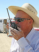 An older gentleman in a wide-brim hat, western shirt and polycarbonate sunglasses holds a 2-way radio to his mouth with a dust-covered hand.