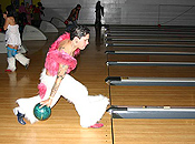 A short-haired woman, wearing a pink fur top and white fur pants shows form and style as she throws her bowling ball.