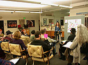 A woman on a stool presents information on a tackboard to a dozen or so attendees.  Photos of fire arts add splashes of color and interest to the meeting room walls.
