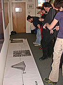 A row of standing people examine a long banner, spread across the floor along a wall.  Images of art projects are laid out along the banner.