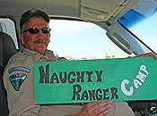 A uniformed BLM ranger seated in his truck holds a sign that states NAUGHTY RANGER CAMP.