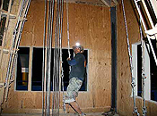 A man pulls down on a steel cable in a windowed room roughed out of plywood. He wears cargo shorts, a long-sleeve shirt and a cowboy hat with a headlamp.