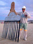 A man in a plastic jumpsuit, hard hat, goggles and gloves holds a giant 'hair pick' approximately 7 feet tall.