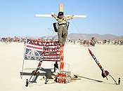 Decorative missles mounted on the ground point at the face of a gagged effigy tied to a cross.  An inverted flag tagged with anarchist graffiti is propped up nearby.