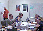 a group of four casually-dressed people work on laptops at a conference table while a fifth person draws on a map on the wall.