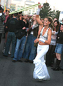 A smiling woman in a silver halter and white pants spins a hoop high in the air around her upstretched hand for a crowd circled around her.