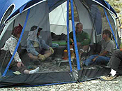 Four campers sit on the desert floor and converse inside a screened tent while a fourth person, also seated, straddles the corner of the structure.