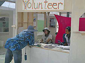 A person leans on the counter of a volunteer booth while speaking to two recruiters seated inside.