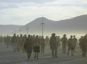 Dozens of Black Rock Rangers walk through blowing dust along a road marked by wooden lamp posts.