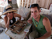 A man pauses from his game of backgammon to smile for the camera as his female opponent smiles patiently under her sunbonnet.