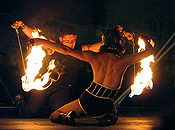 A man and woman kneel, facing each other, with burning poi hanging from their arms, stretched out to their sides.