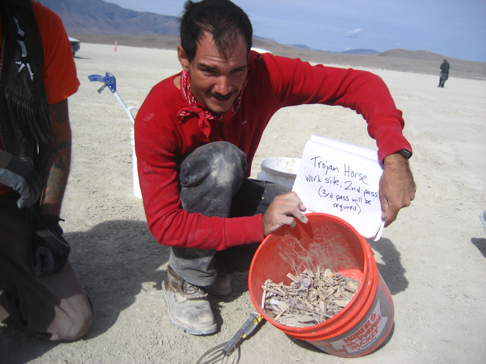 man on playa tipping a full bucket of woodchip MOOP debris towards the camera along with a notebook page that says "Trojan Horse work site, second pass (third pass will be required)