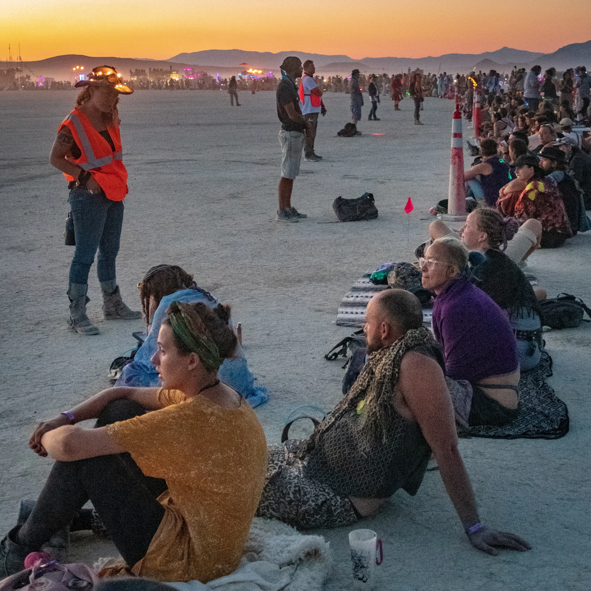 A semicircle of people seated on the ground with a few people in orange vests standing in front of the crowd. 
