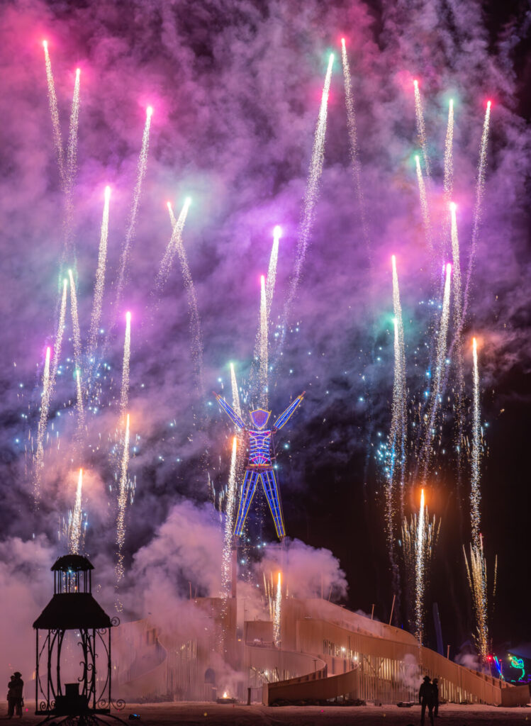 Fireworks shoot into the sky in front of the lighted Man effigy with upraised arms.