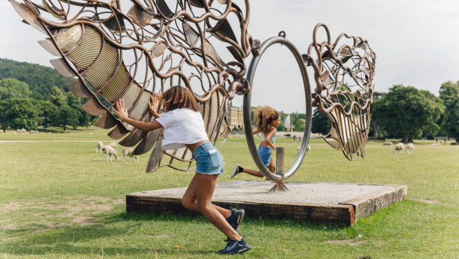 Children play with an interactive metal wing sculpture on a grassy field in England. 