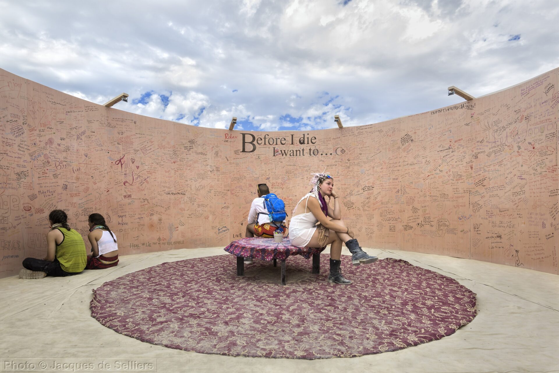 People sit on a bench at the edge of a circular rug in front of a wooden wall that says, "Before I die I want to" and hundreds of statements written by individuals.
