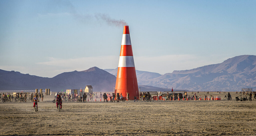 A 60-foot tall wooden traffic cone sculpture on playa with smoke coming out the tip in daylight.