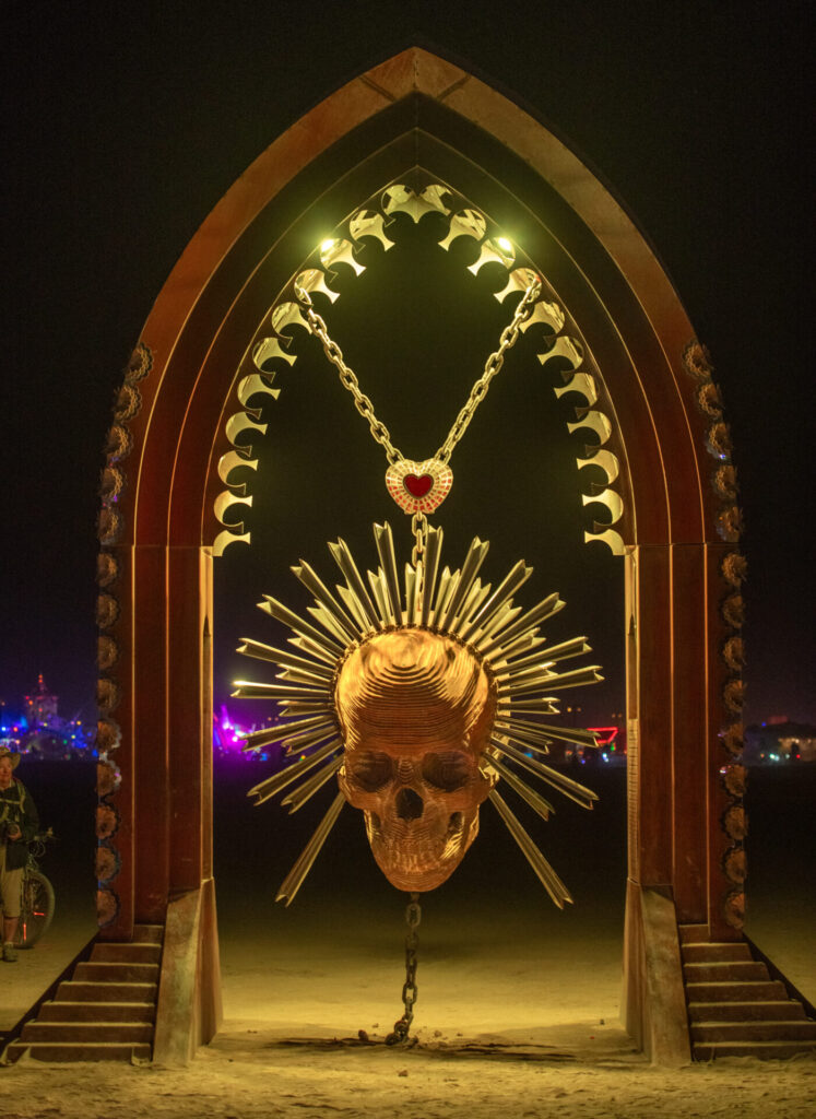 A large-scale sculpture lit in yellow light on playa at night of a skull with a crown of radiating lines from the head suspended from an archway by a y-shaped gold chain with a heart at the center of the y. 