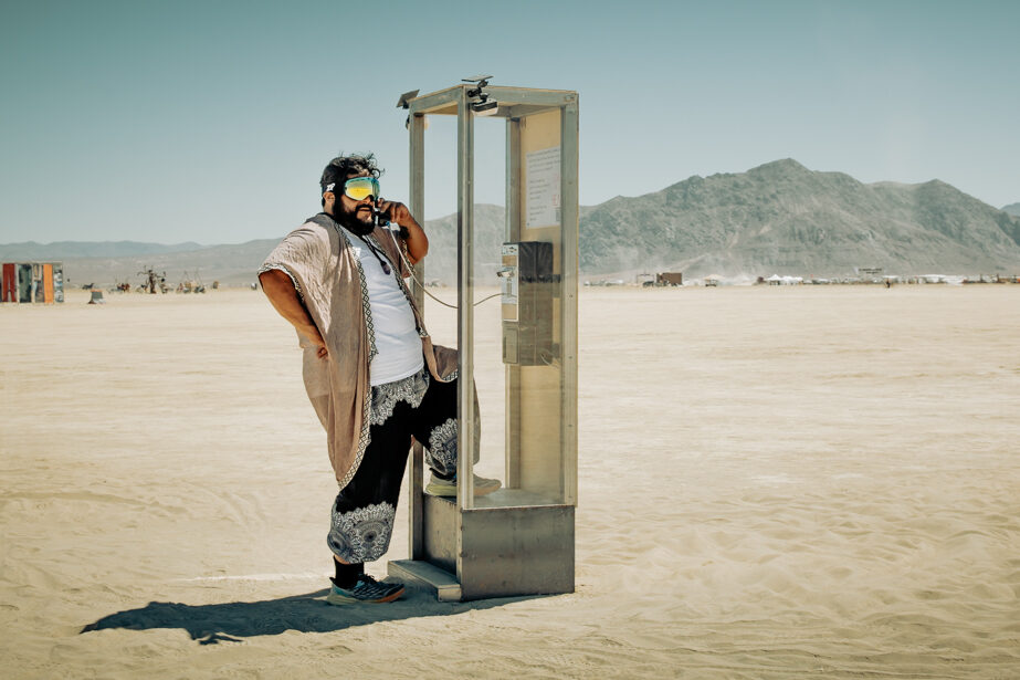 Person standing at phone booth on playa with receiver up to their ear.