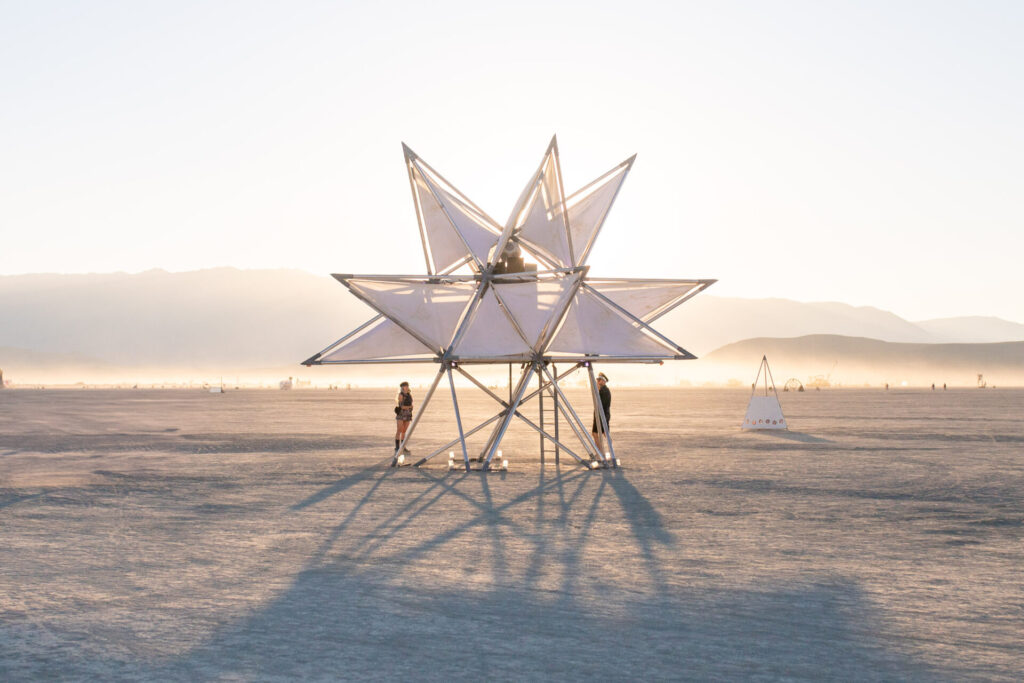 Elevated spiky white art star sculpture on playa with two people looking up at it in daylight.