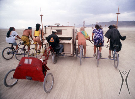 A mutant vehicle with a piano in the middle, attached piano stool on a wheel and three bicycles on each side being ridden along a BRC road. 