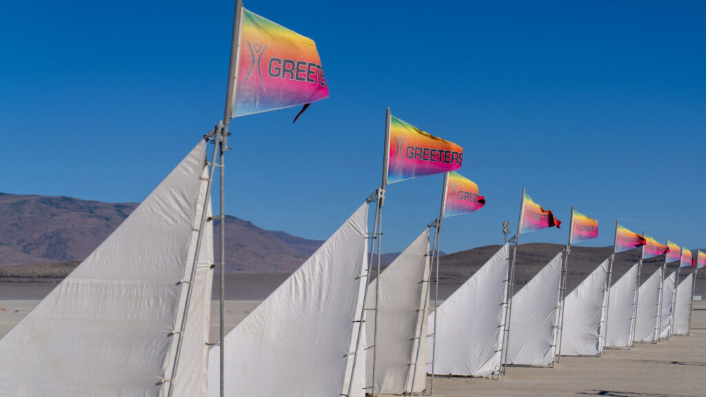 Rows of vibrant Greeter flags and white lanes mark the Greeters stations, where friendly volunteers welcome newcomers to the playa, setting the tone for a unique journey into community and creativity.