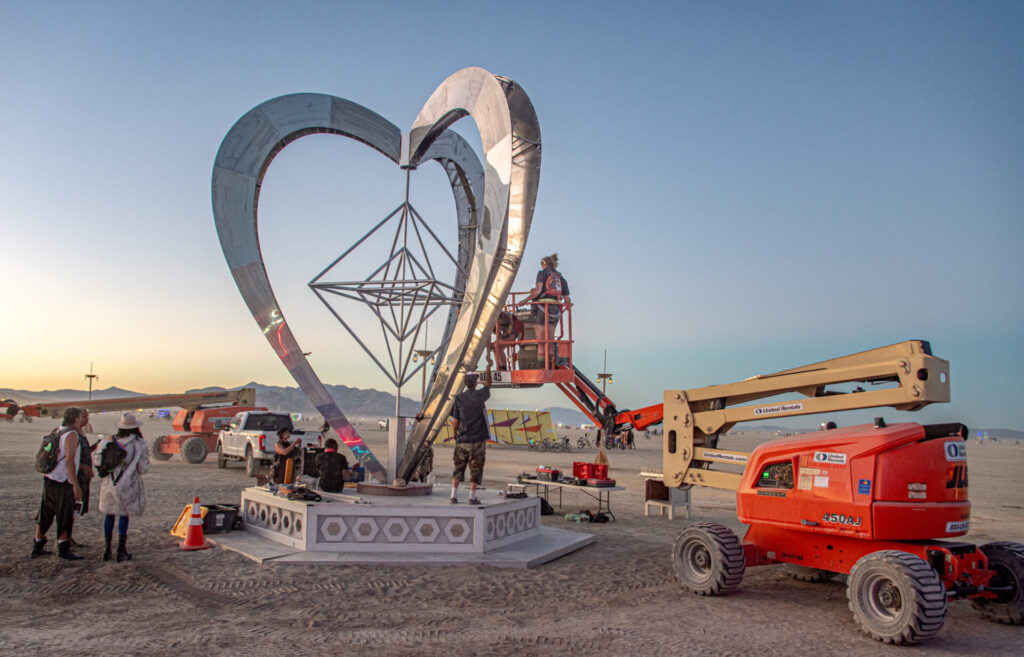 large metal heart sculpture being constructed with a piece of heavy machinery on playa 