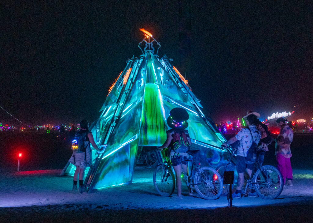 People straddling bikes admiring an  pyramid-like sculpture on playa lit in teal LED lights.