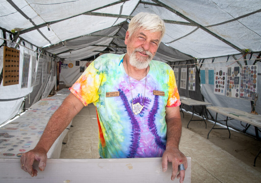 Man in tie-dye shirt with burning man symbol in front of tables full of items made by participants as gifts.