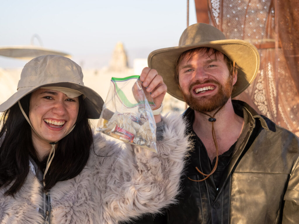 Two burners, a white man and woman, hold up a plastic bag filled with trash found on playa aka MOOP.