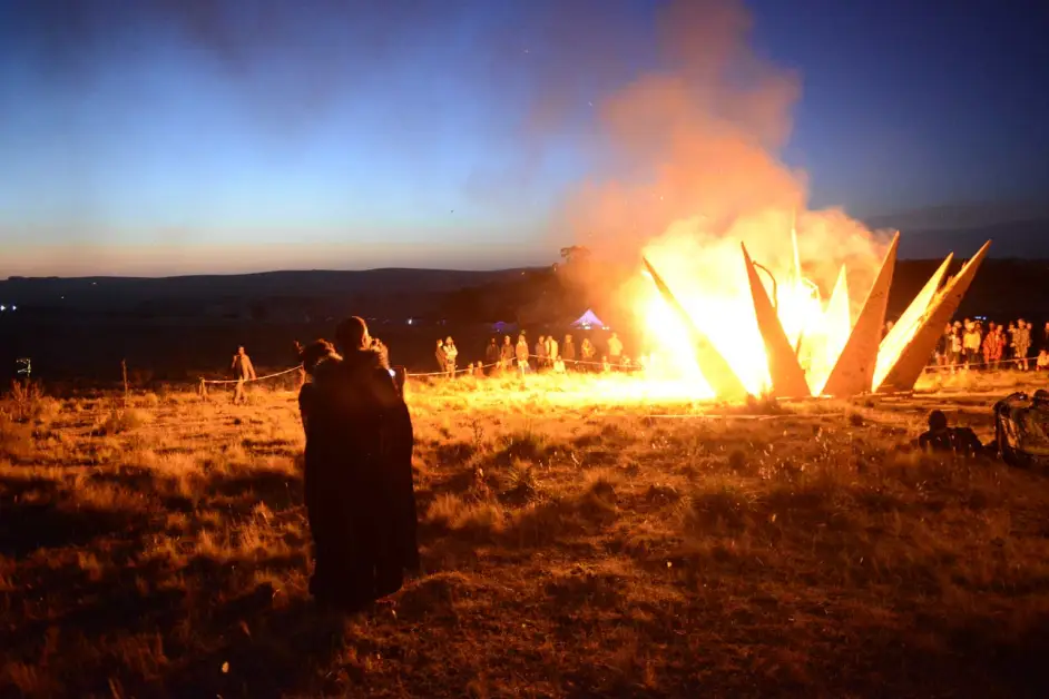 Bonfire burning in a field with people standing around it at a distance.