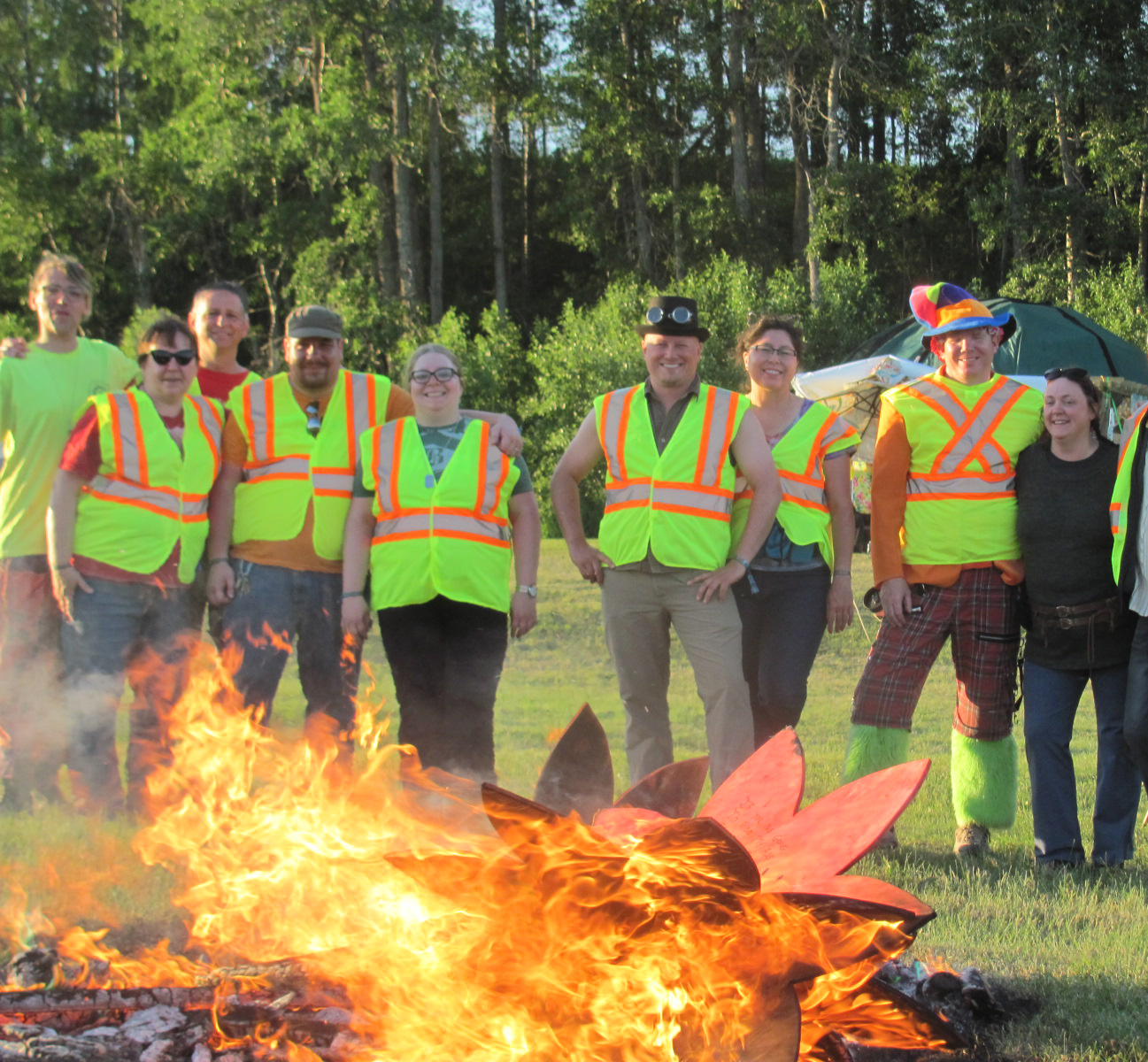 A group of people in safety vests stand near a red wooden flower on fire.