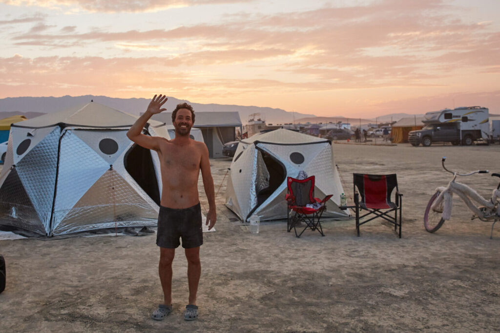 A man waves at the camera with shift pods and the sunset behind him