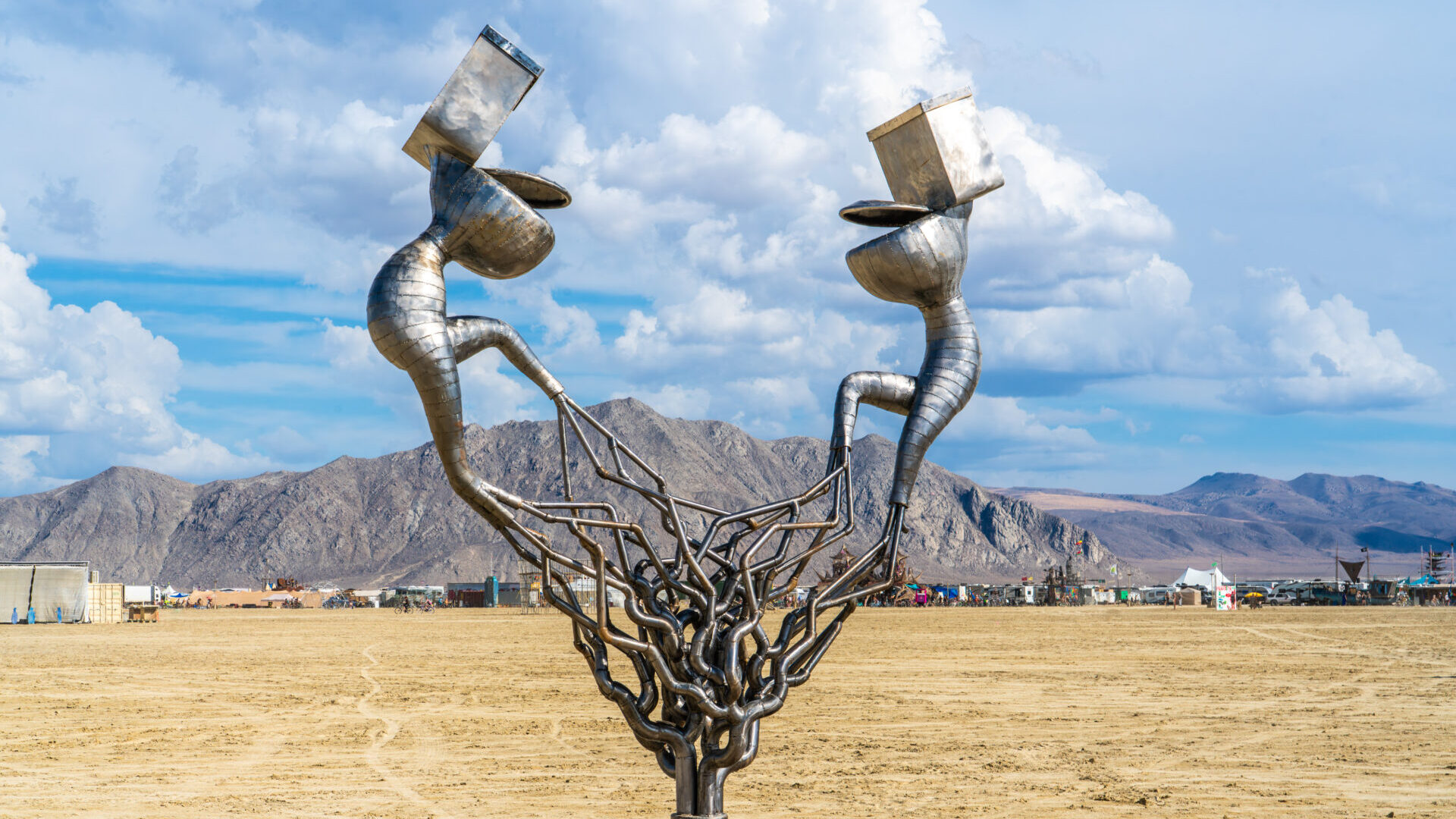 Welded metal sculpture of two toilets facing each other with seats partly open. The toilets are attached to humanoid lower torso and legs that come together in a web of metal pipes to a single pipe at the base.