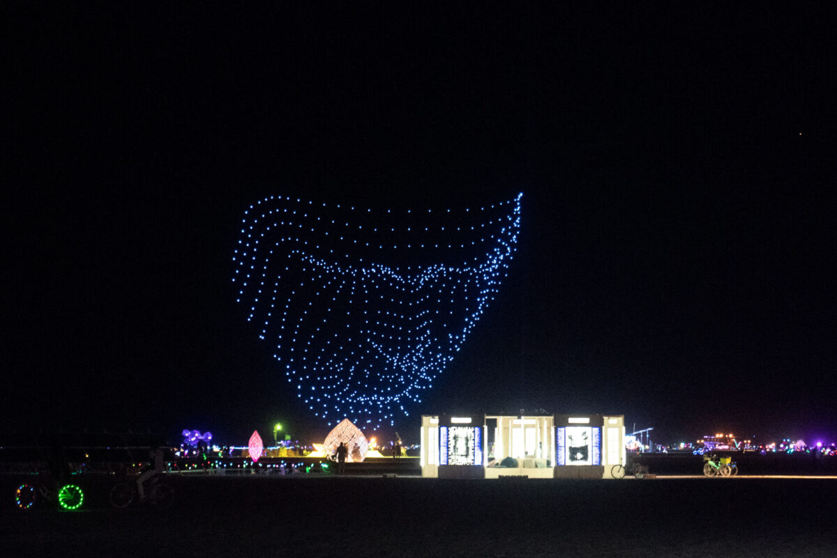 A swarm of drones form the shape of a face looking down onto the playa.