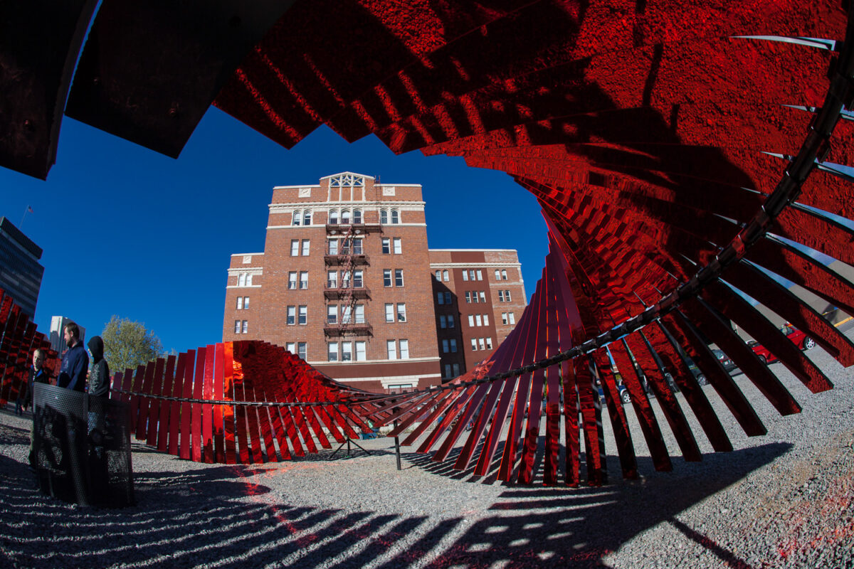 Forming a complete circle, this sculpture is made of metal rods and planks. Standing outside the sculpture, you can see the shape the planks make as they are slanted one a little more than the next. Standing inside the sculpture however, is where the true magic of this work of art lies. Inside of the sculpture the planks are a reflective red surface meant to represent blood as the entire sculpture as a whole represents human DNA. The red planks reflect back on the viewer their world around them so as to show them their connection to the people in their surroundings.
