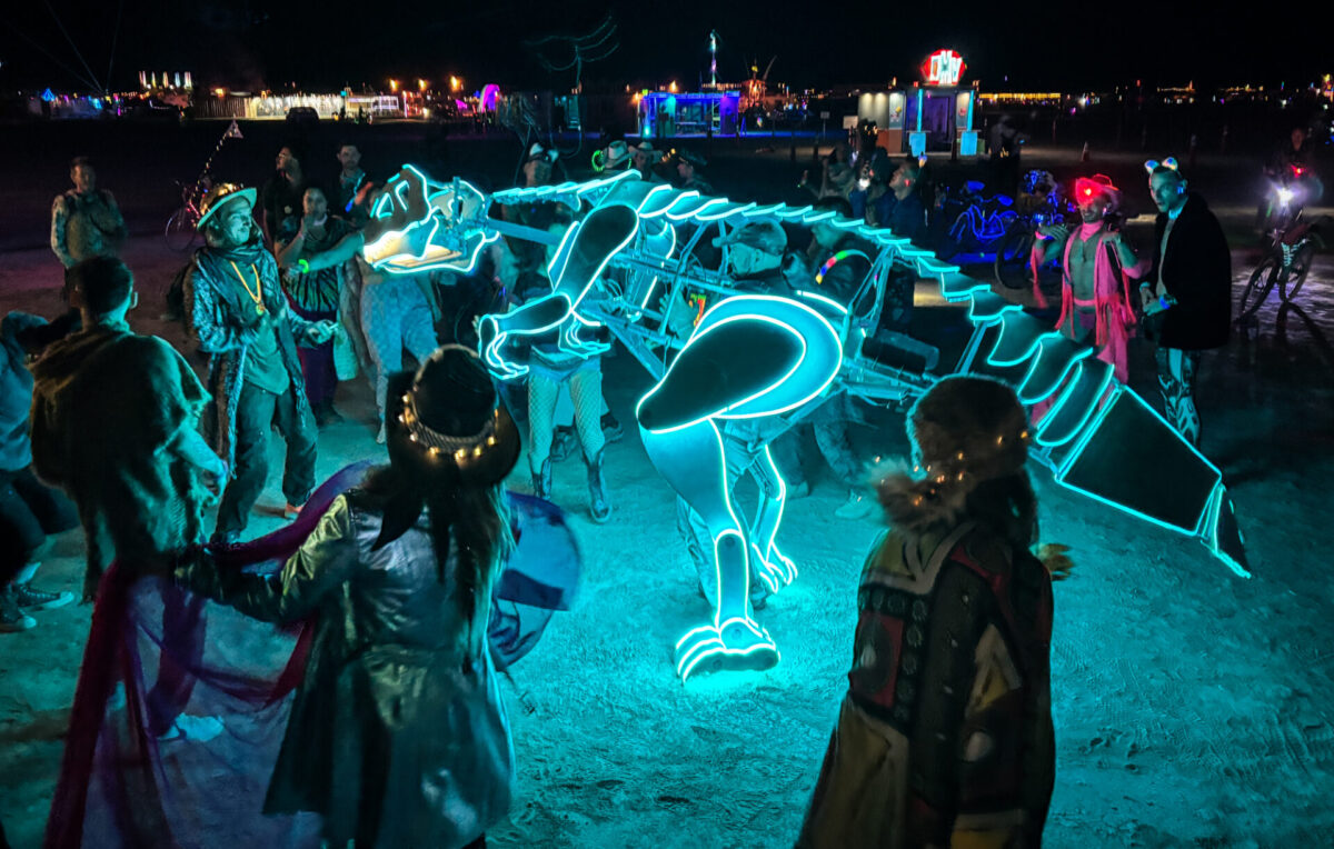 Person wearing a mechanical dinosaur costume trimmed in green neon dancing in a crowd
