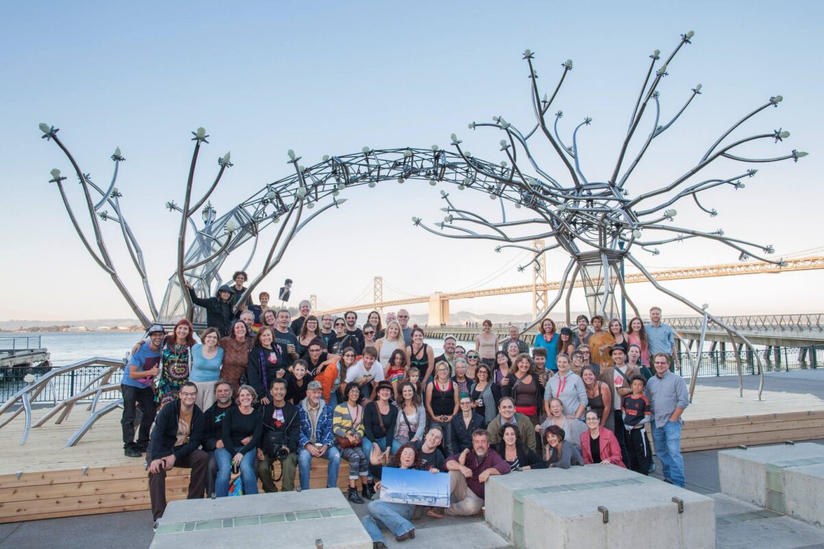 A massive somatic nerve made of metal in front of the Bay Bridge with a group of artists under it.