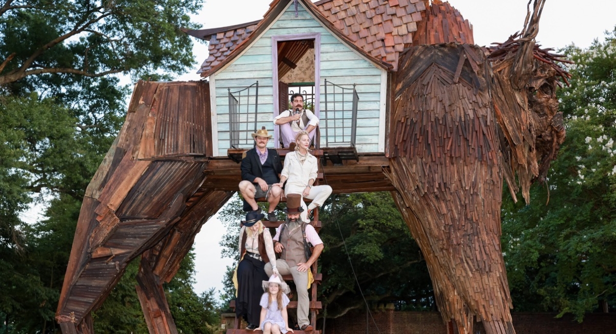 Artists and family members sitting on the ladder steps to art piece "Burden of the Beast," a tall beast made of wood pieces with a tiny house at the center top