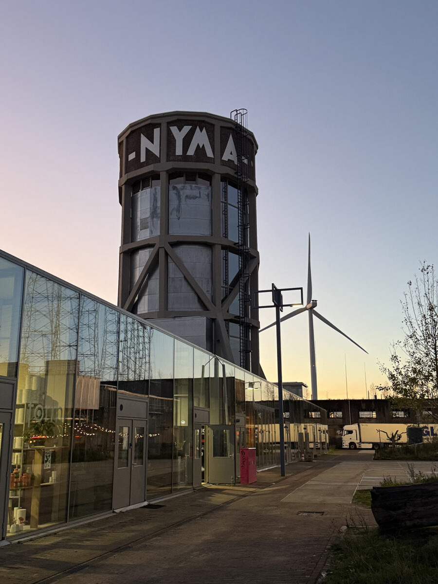 A glass-walled building with a cylindrical tower that says, "NYMA" and windmill in the background