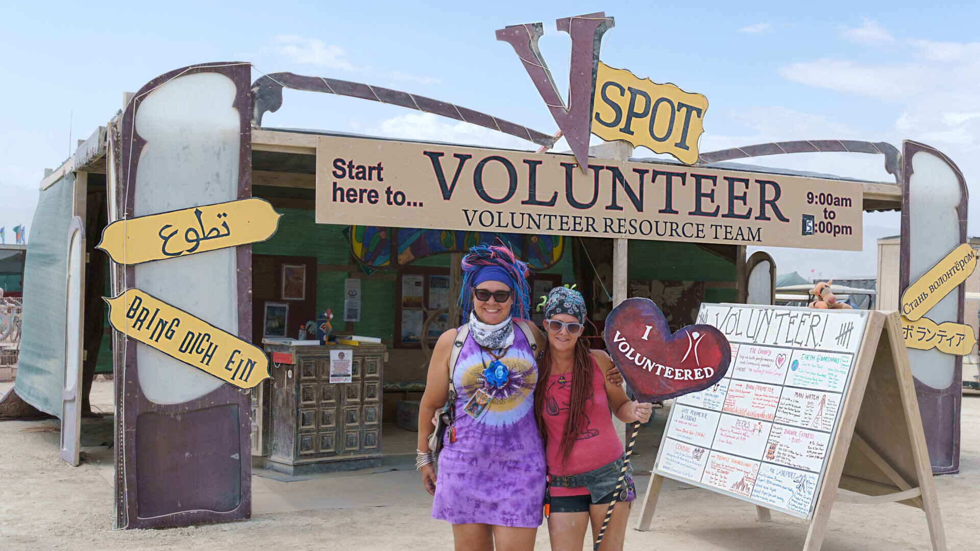 Two volunteers smile and wave at the V Spot, Volunteer headquarters in Black Rock City.
