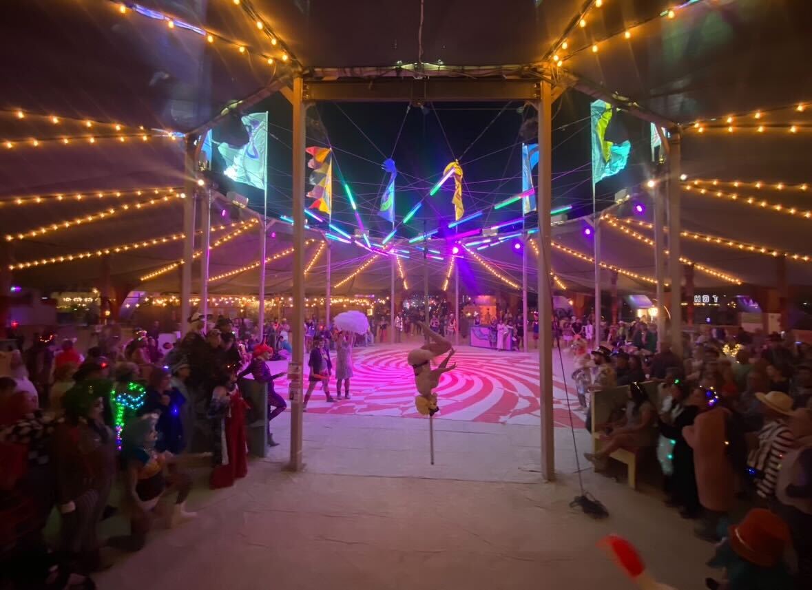 Nighttime aerialist and acrobatic performance in the Center Camp Canopy with multiple performers and large seated audience.