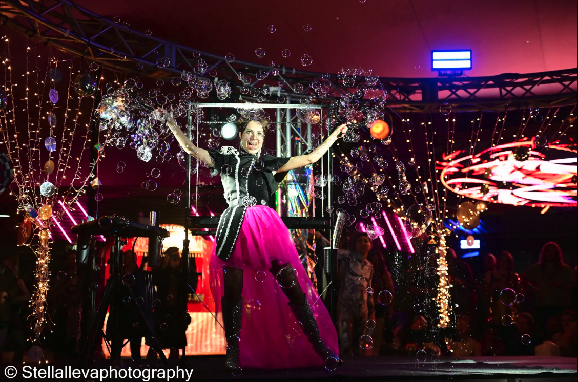 A woman in a black shirt and pink tutu stands on stage amongst cascading bubbles.