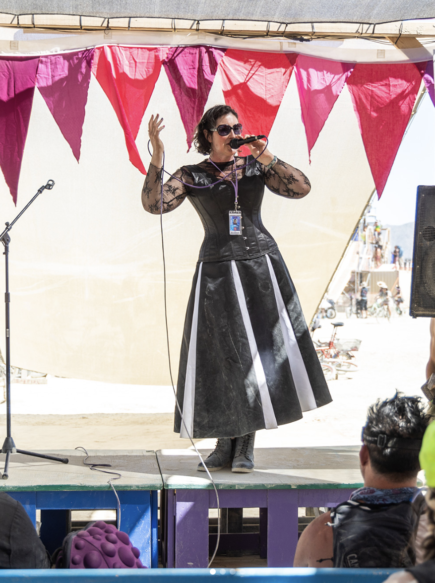 A person standing on a stage speaks into a microphone with an arm raised emphatically.
