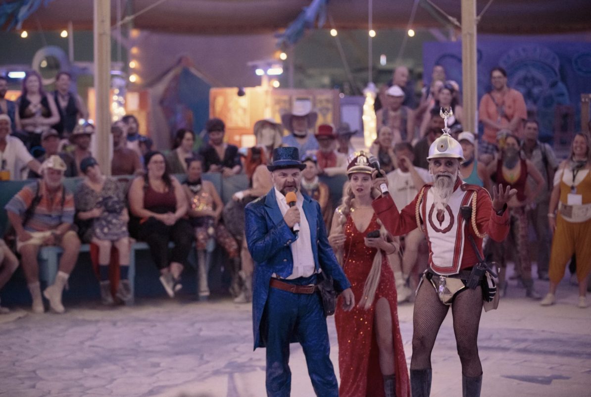 Three people with microphones stand at the center of a ring of people, the masters of ceremony for an event.