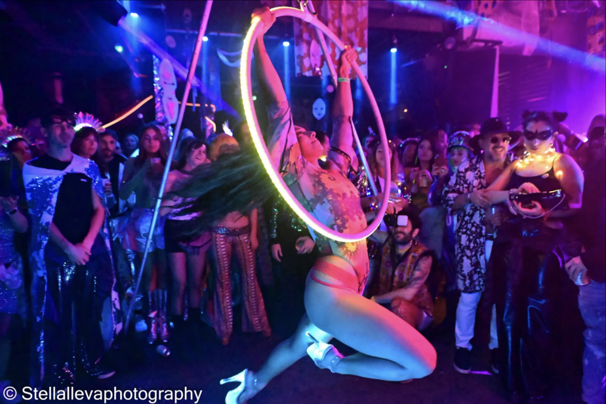 Woman in bodysuit and silver high heels swings from a suspended hula hoop in a nightclub setting while festively dressed Burners look on.
