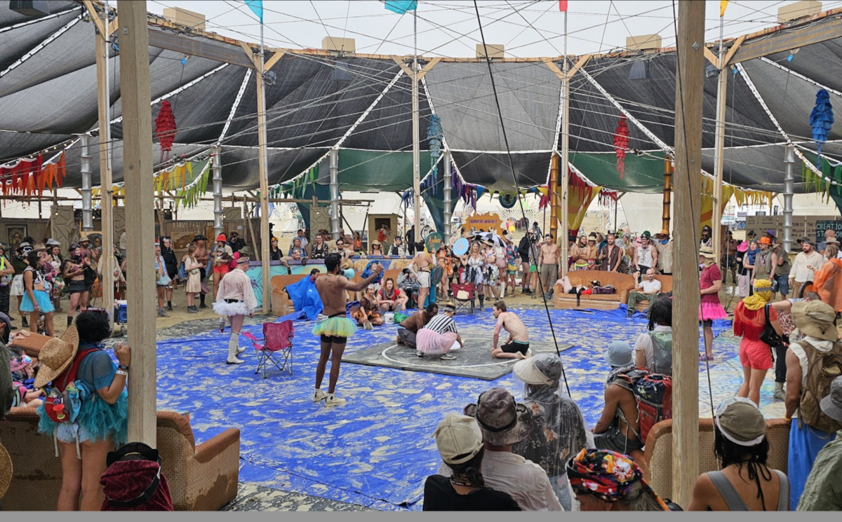 People stand in a circle around a regulation-size wrestling ring while a referee kneels with wrestlers and announcer speaks into a microphone. 