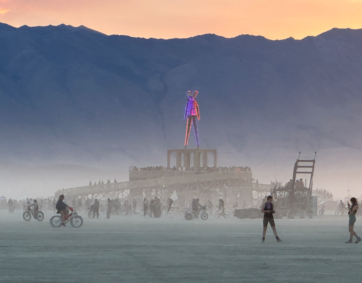People walk and bicycle around the Burning Man Pavilion, with the effigy sculpture on top lit in orange and purple neon, at sunset.