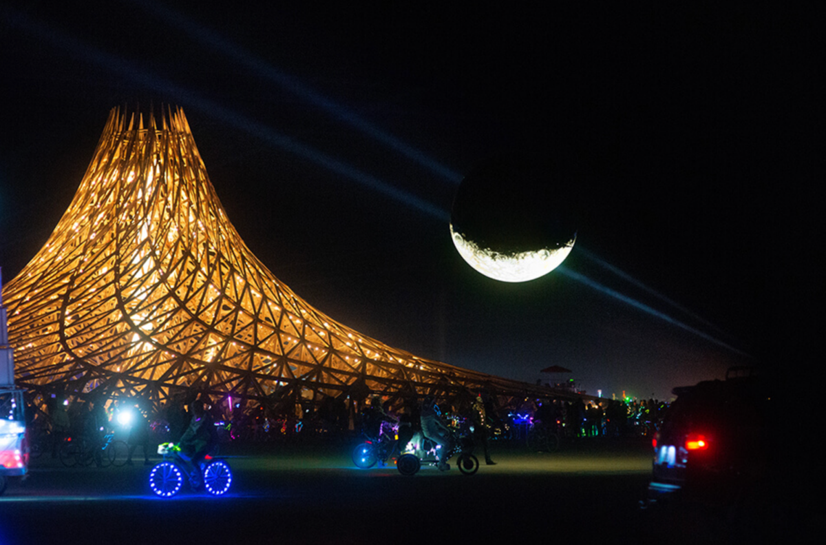 A large moon sculpture floats above the playa at night.